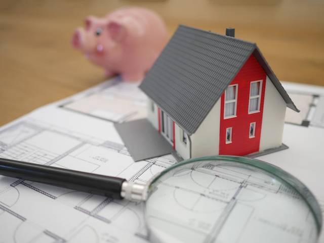Una casa en miniatura sobre unos planos, con una lupa delante y una hucha con forma de cerdo al fondo