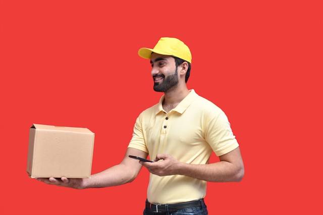 Un repartidor con gorra y polo amarillos sostiene una caja de cartón en una mano y un móvil en la otra, sobre fondo rojo