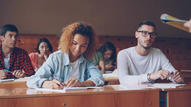 Estudiantes en los bancos de un aula universitaria: una joven toma apuntes y un chico con gafas mira al frente