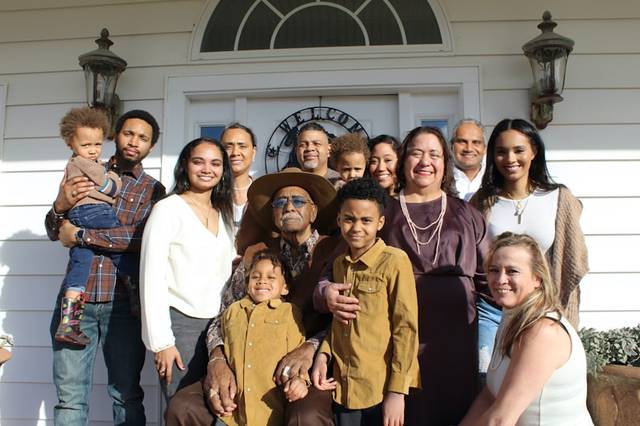 Una familia numerosa de varias generaciones posa sonriente ante la puerta de una casa blanca