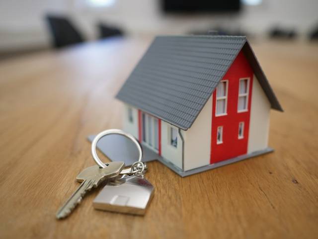Una casa en miniatura, blanca y roja, junto a un llavero con una llave, sobre una mesa de madera