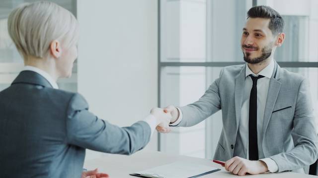 Un hombre con traje gris y una mujer de pelo corto se dan la mano por encima de una mesa con documentos